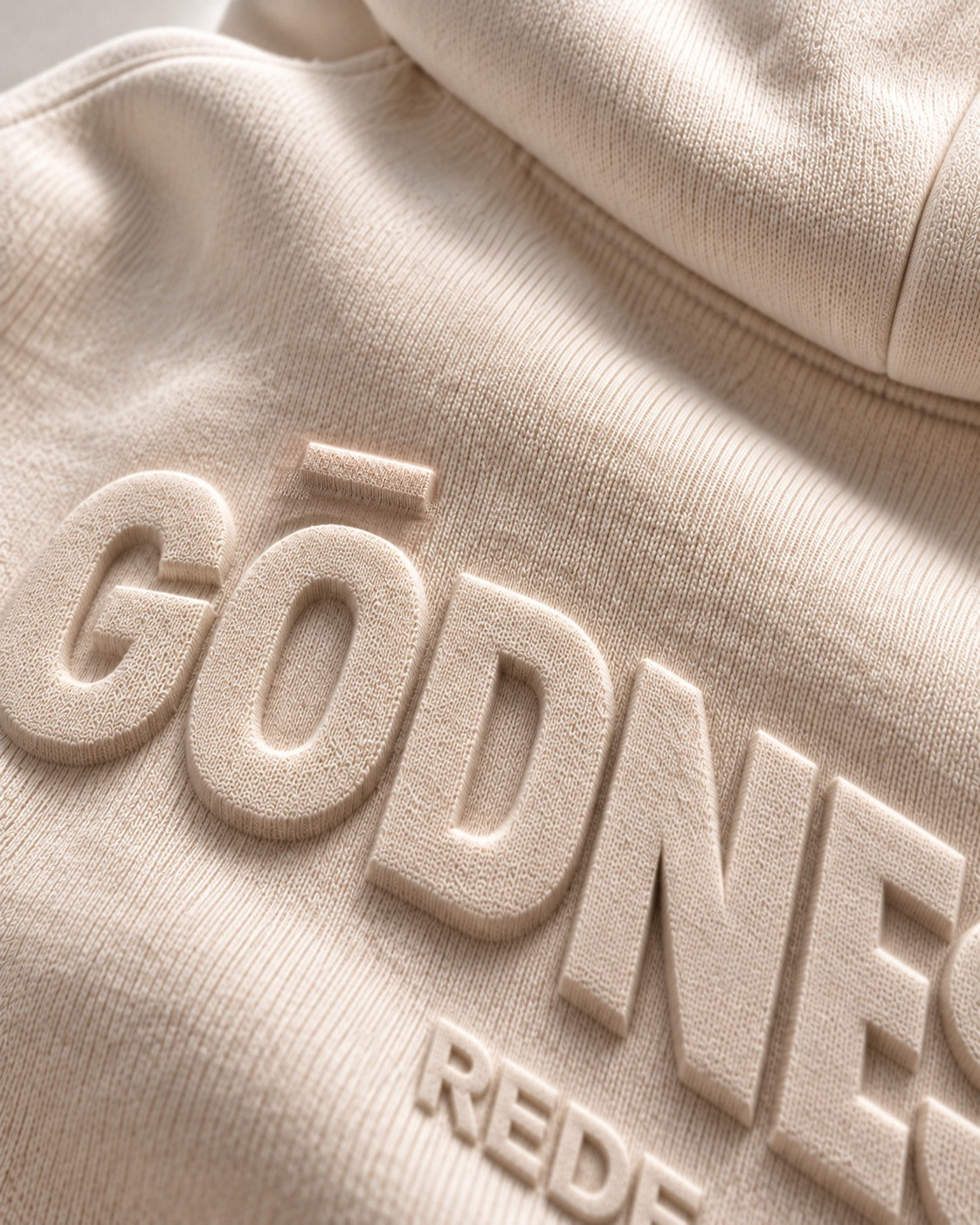 Teaser concept: angled light defines raised tonal lettering in a close-up of bone hoodie fleece.
