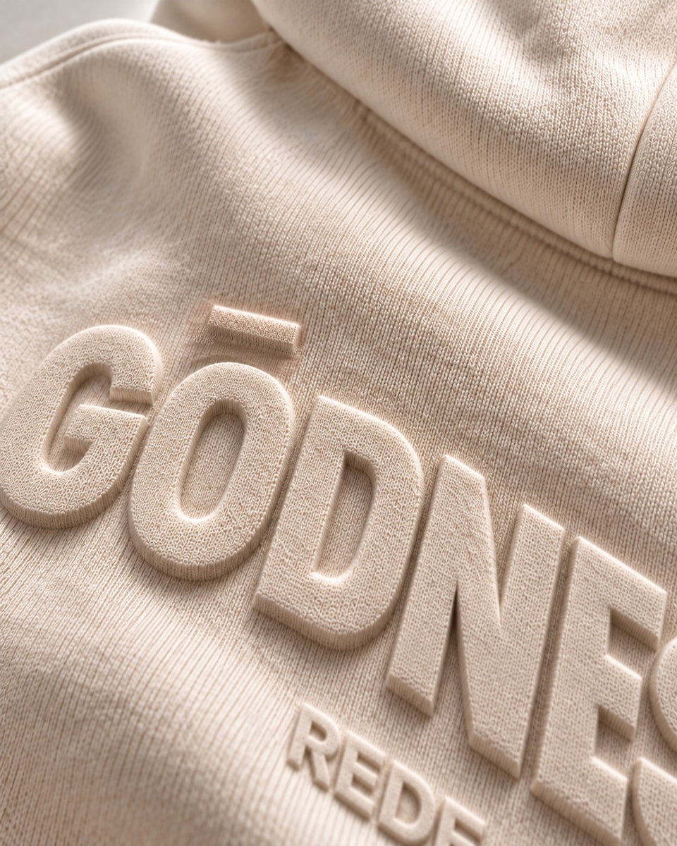 Teaser concept: angled light defines raised tonal lettering in a close-up of bone hoodie fleece.